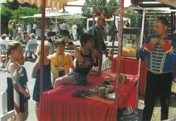 Ein Mann in einem roten Weste und ein Mann mit Muskeln sitzen an einem Tisch mit Snacks, während eine Frau mit einer Lanyard im Hintergrund steht. Im Hintergrund sind Menschen und ein Stand zu sehen.