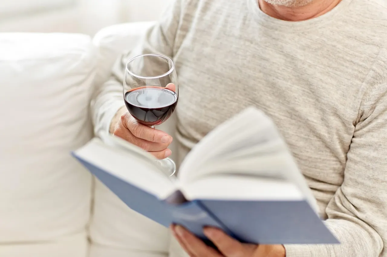 Eine Person hält ein Glas Rotwein und ein aufgeschlagenes Buch.