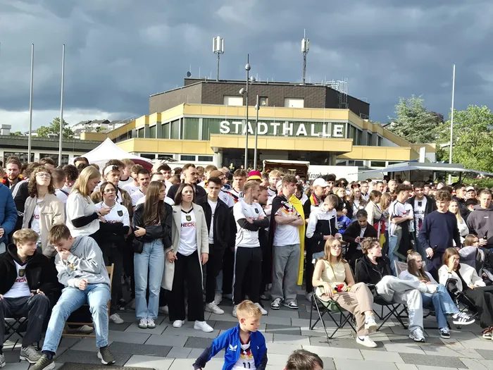Große Menschenmenge vor der Stadthalle, viele tragen Fußballtrikots und schauen in eine Richtung, vermutlich ein Bildschirm. Im Hintergrund sind Antennen zu sehen und ein bewölkter Himmel.