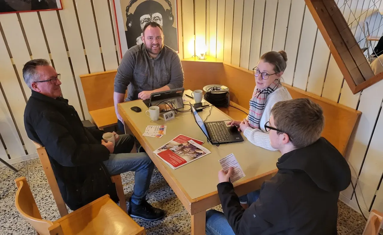 Vier Personen sitzen an einem Holztisch in einem hellen Raum zusammen, zwei Laptops und Broschüren liegen darauf. Sie unterhalten sich und schauen auf Notizen oder Bildschirme.