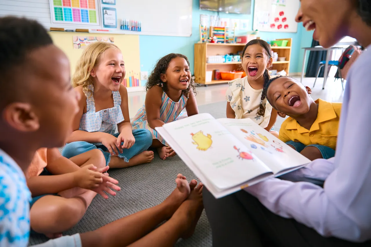 Eine Lehrerin liest einer Gruppe lachender Kinder in einem Klassenzimmer aus einem großen Bilderbuch vor.