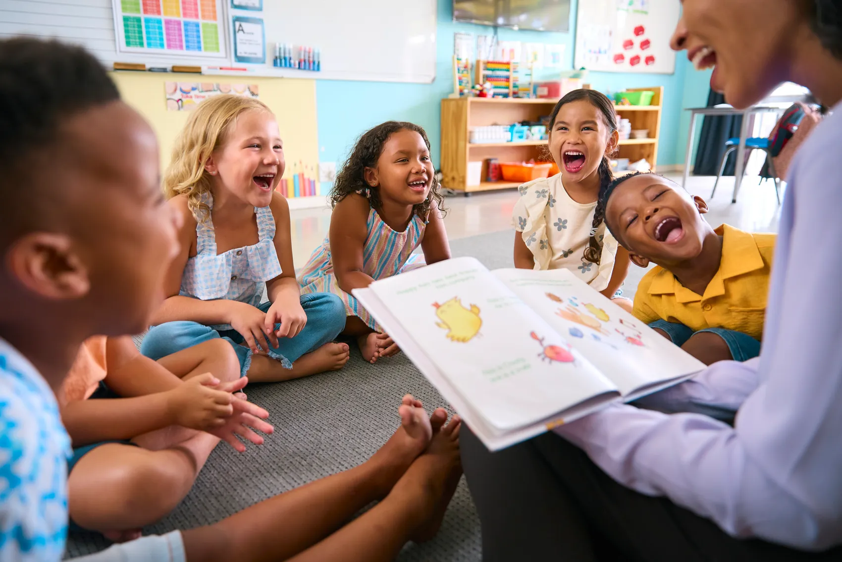 Eine Lehrerin liest einer Gruppe lachender Kinder in einem Klassenzimmer aus einem großen Bilderbuch vor.