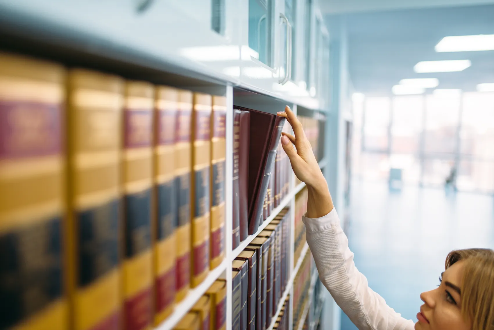 Eine Frau greift in einem Archiv nach einem Buch in einem Regal. Die Regale sind voll mit Büchern in verschiedenen Farben.