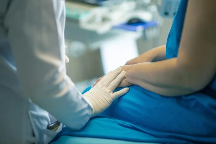 Ein Arzt oder eine Ärztin mit weißen Handschuhen hält die Hand einer Patientin, die auf einer Liege sitzt.