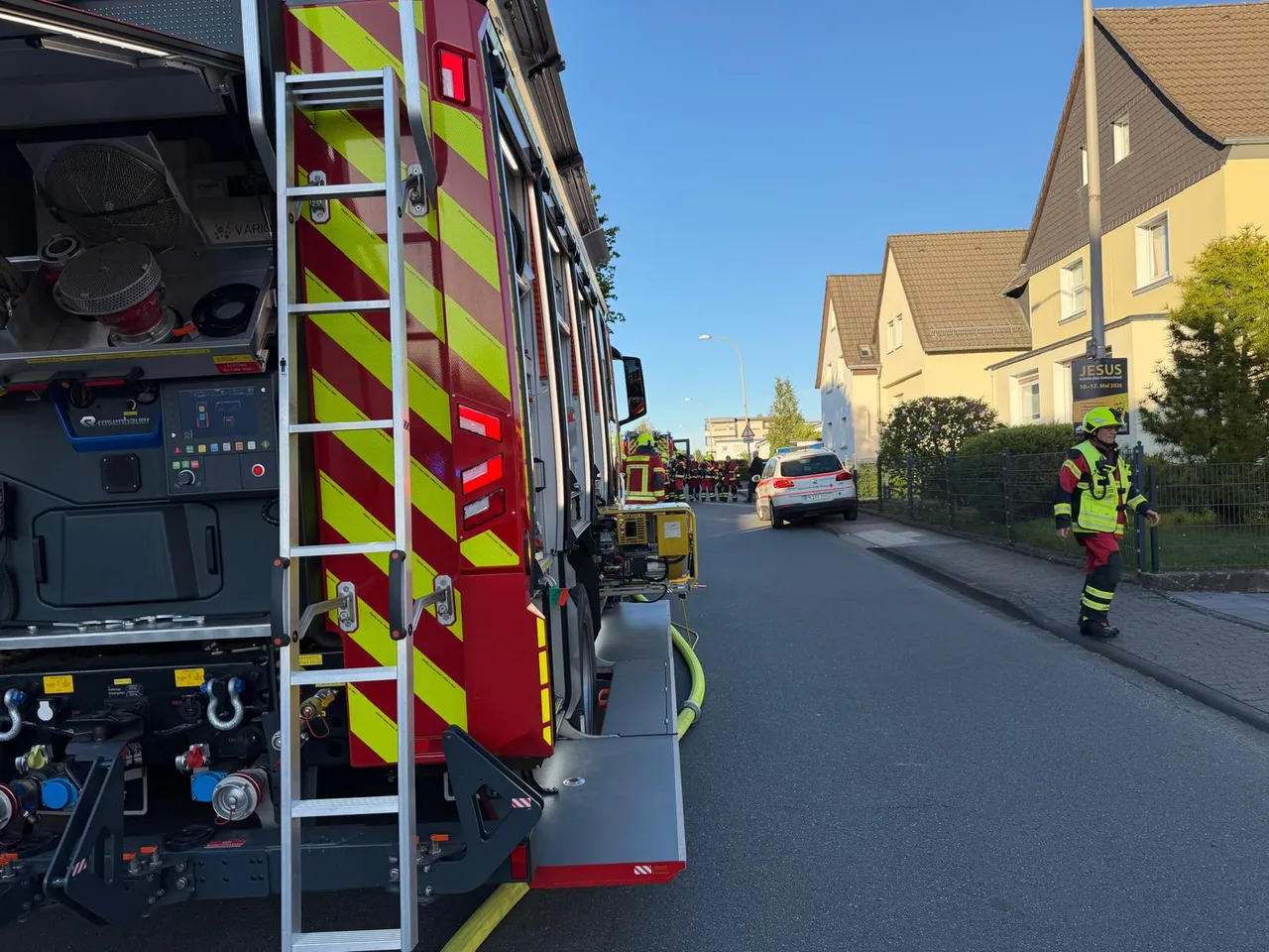 Roter Feuerwehrauto mit ausgefahrener Leiter steht an einer Straße, während Feuerwehrleute einen Brand in einem Wohngebiet bekämpfen.