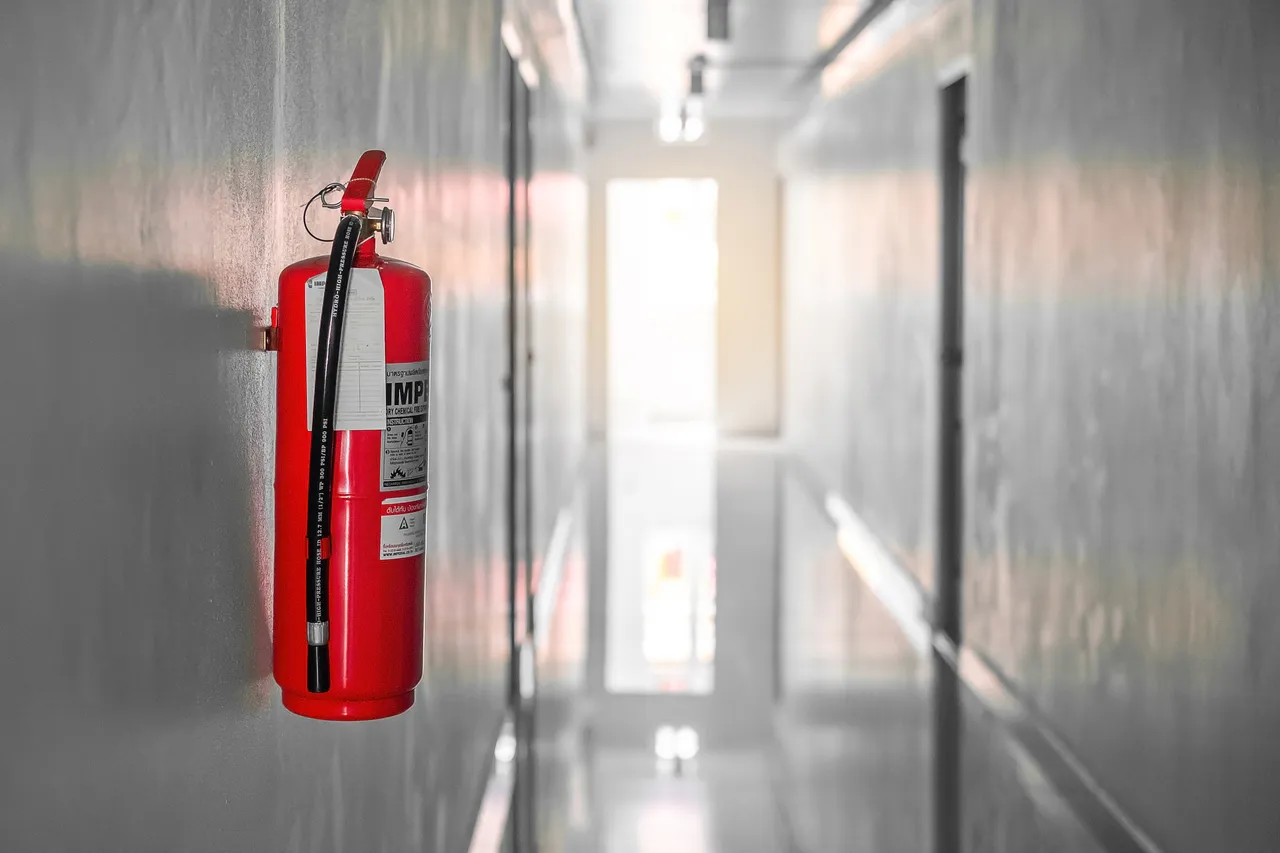Ein roter Feuerlöscher mit Manometer hängt an einer grauen Wand in einem hellen Flur.