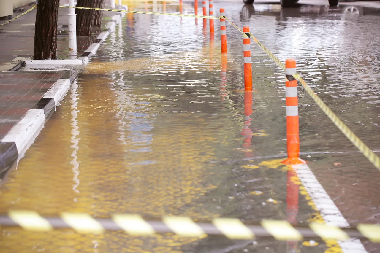 Eine überflutete Straße mit orangefarbenen Absperrkegeln und gelb-schwarzem Warnband.