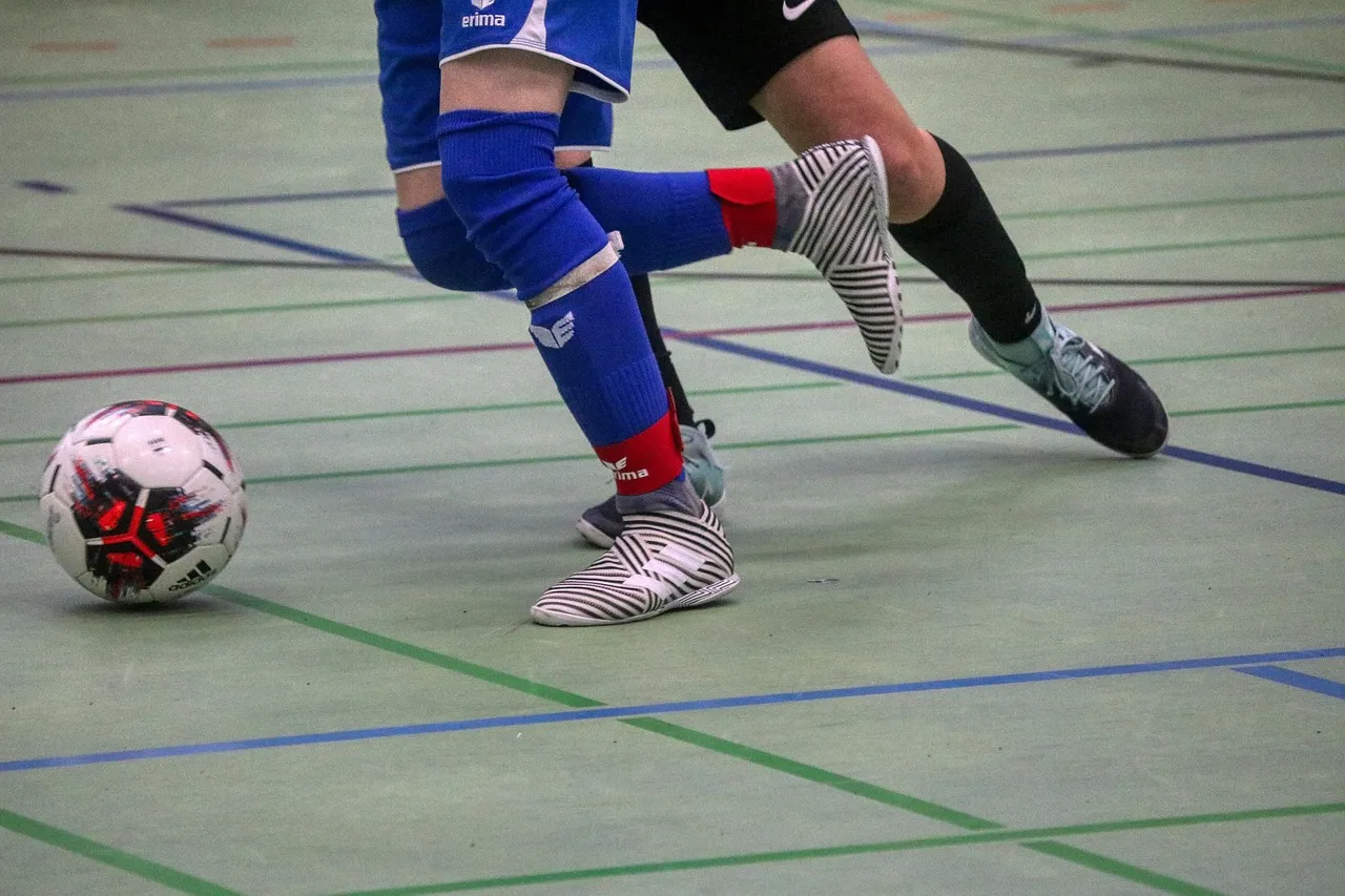 Das Bild zeigt die Beine von zwei Fußballspielern und einen Fußball auf einem Hallenfußballfeld. Ein Spieler trägt blaue Shorts und Stutzen mit roten Akzenten und schwarz-weiße Schuhe, der andere Spieler trägt schwarze Shorts und graue Schuhe.