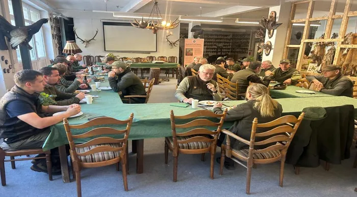 Eine Gruppe von Männern sitzt an langen, grün gedeckten Tischen in einem Raum mit Jagdtrophäen an den Wänden.