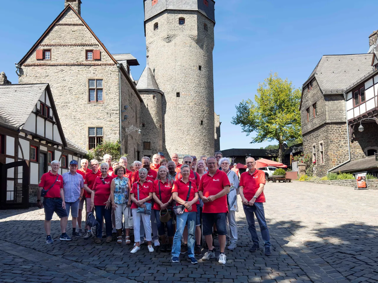 Eine Gruppe von Menschen in roten T-Shirts posiert auf dem gepflasterten Innenhof der Burg Altena vor einem hohen Steinturm und Fachwerkhäusern.