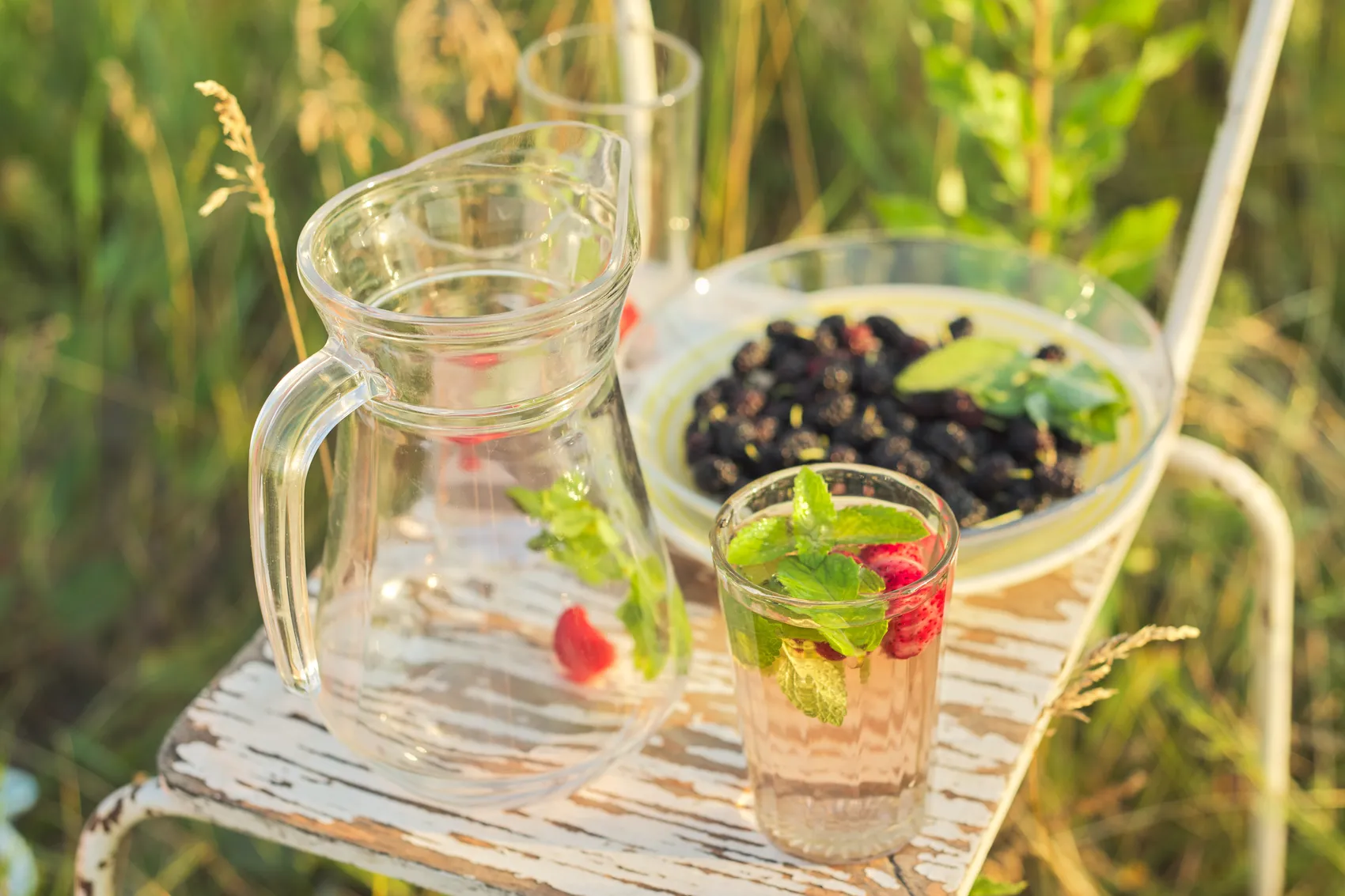 Ein Glas und ein Krug mit einem Getränk, verziert mit Minze und Erdbeeren, stehen neben einer Schüssel mit Brombeeren auf einem weißen Tisch im Freien. Im Hintergrund ist grünes Gras zu sehen.