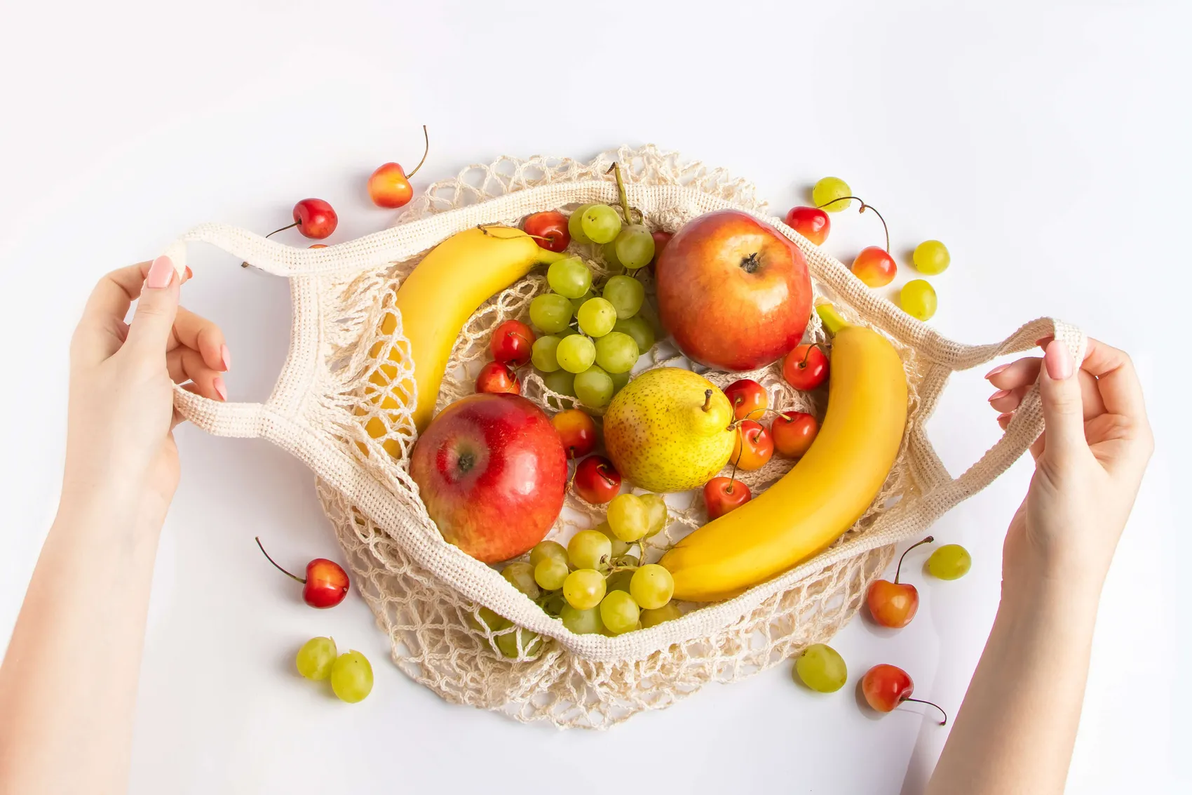 Eine Hand hält eine offene, beige Netztasche gefüllt mit Äpfeln, Birnen, Bananen, Trauben und Kirschen.