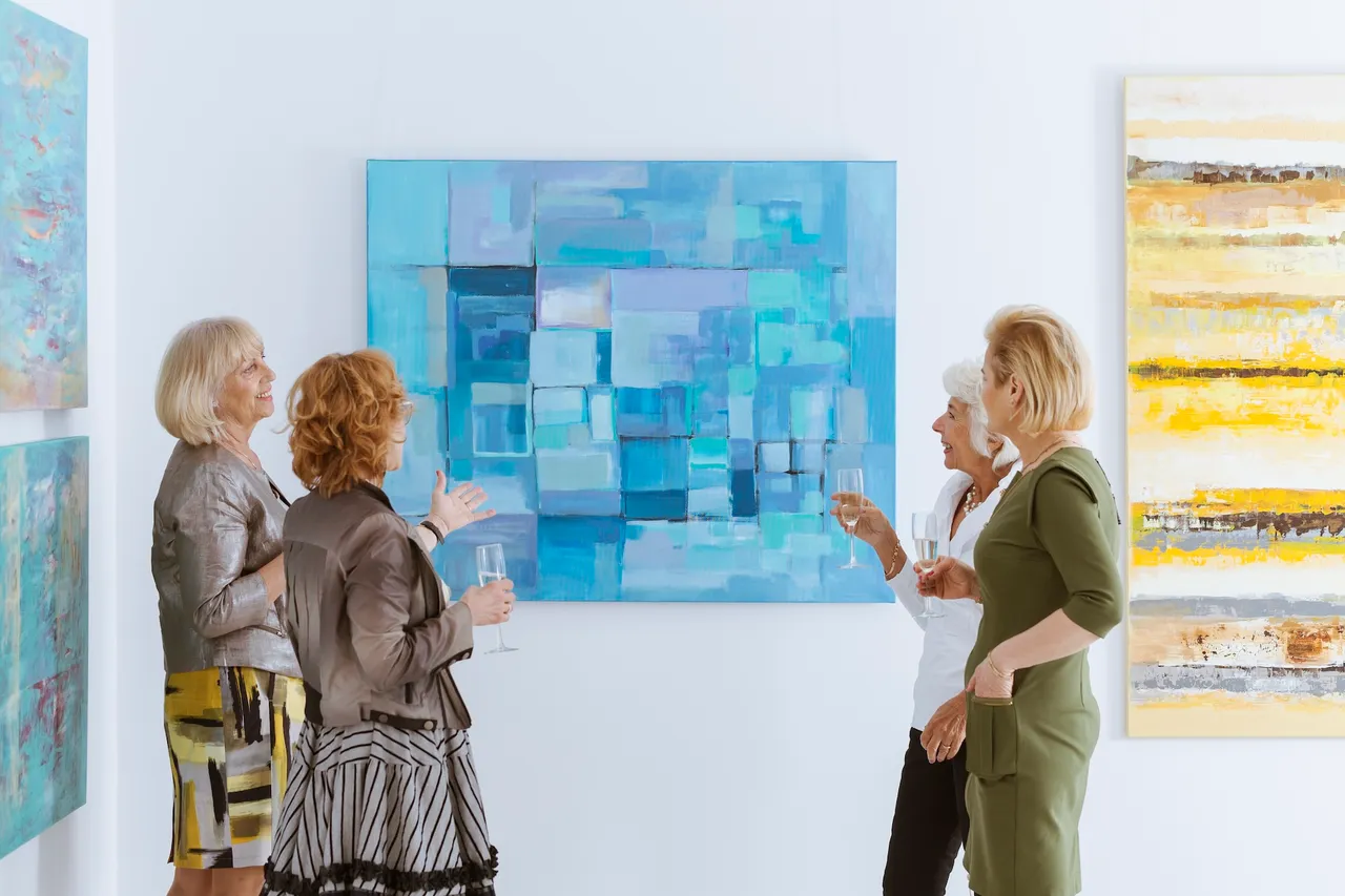 Vier Frauen mittleren Alters betrachten ein abstraktes Gemälde in hellen Blautönen während einer Vernissage in einer Galerie.