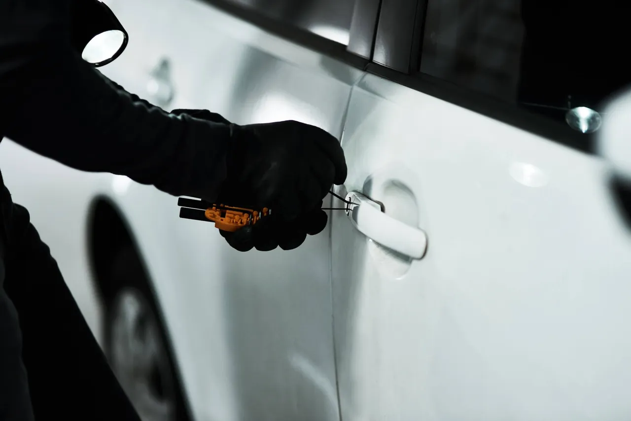 Eine Person in schwarzen Handschuhen manipuliert mit einem Werkzeug den Türgriff eines weißen Autos bei Nacht.