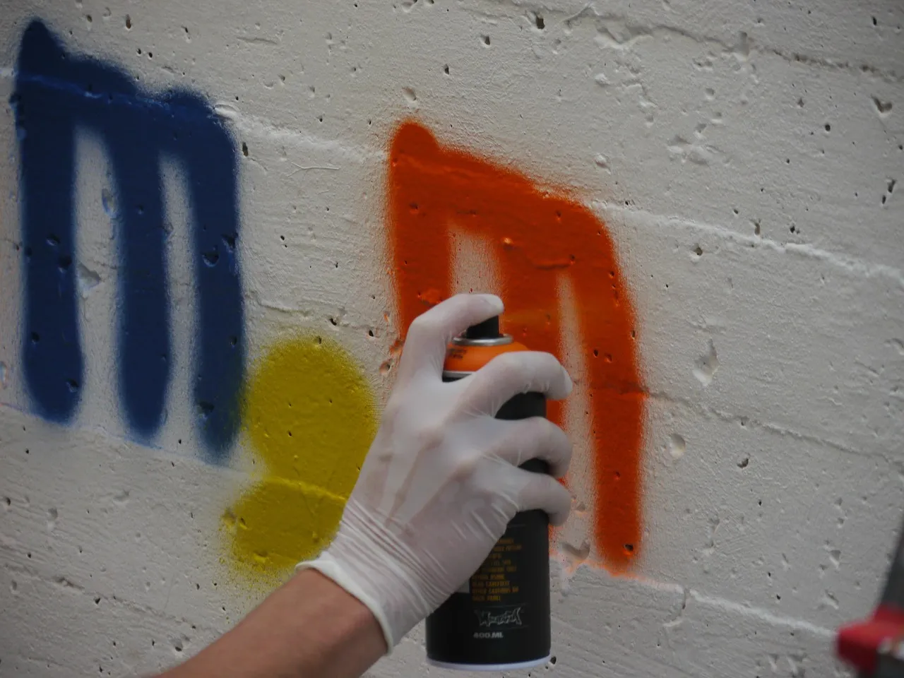 Eine Hand mit weißem Handschuh sprüht orangefarbene Farbe auf eine weiße Wand neben blauen und gelben Graffiti-Elementen.