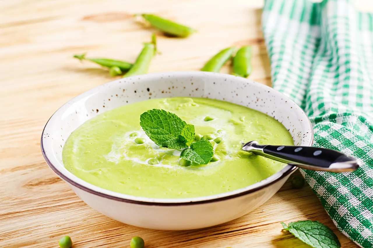 Eine Schüssel cremige, grüne Erbsensuppe mit einem Minzblatt und einem Löffel darin, umgeben von frischen Erbsen und einem karierten Küchentuch auf einem Holztisch.