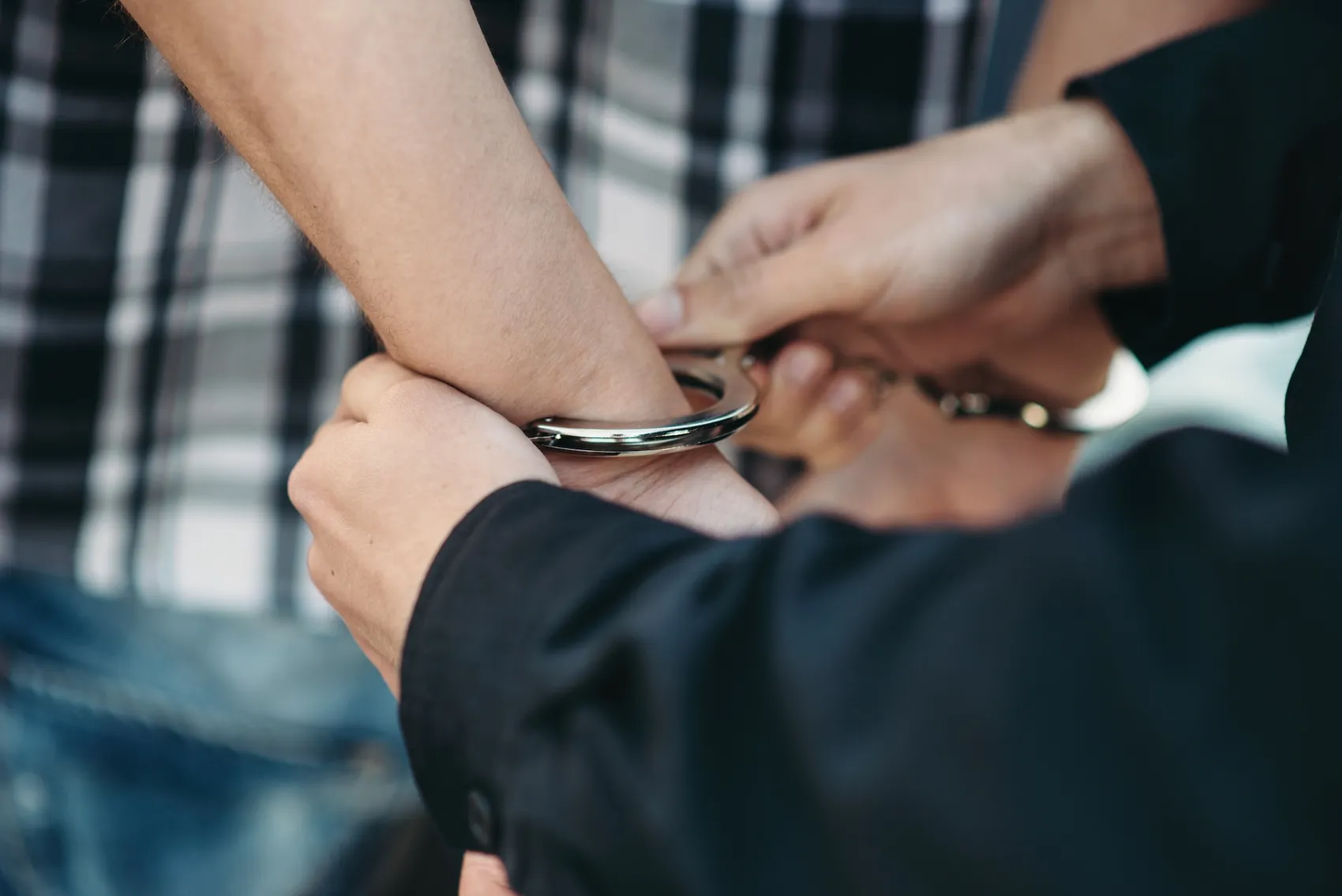 Handschellen werden einer Person angelegt, die ein kariertes Hemd trägt. Die Aufnahme ist aus der Nähe und zeigt die Handgelenke und die Hände des Polizisten.
