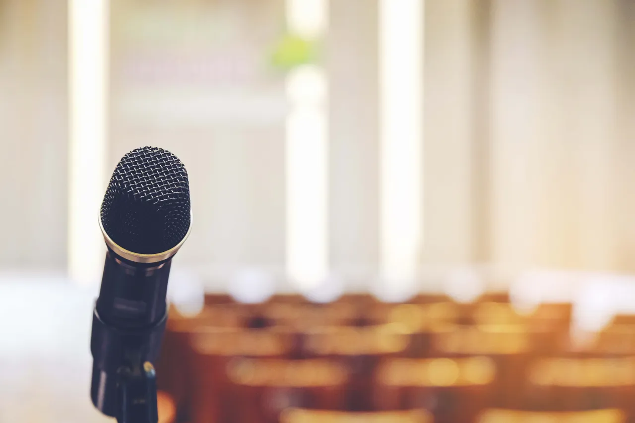 Nahaufnahme eines schwarzen Handmikrofons vor einer verschwommenen Reihe von Sitzen in einem Konferenzraum.