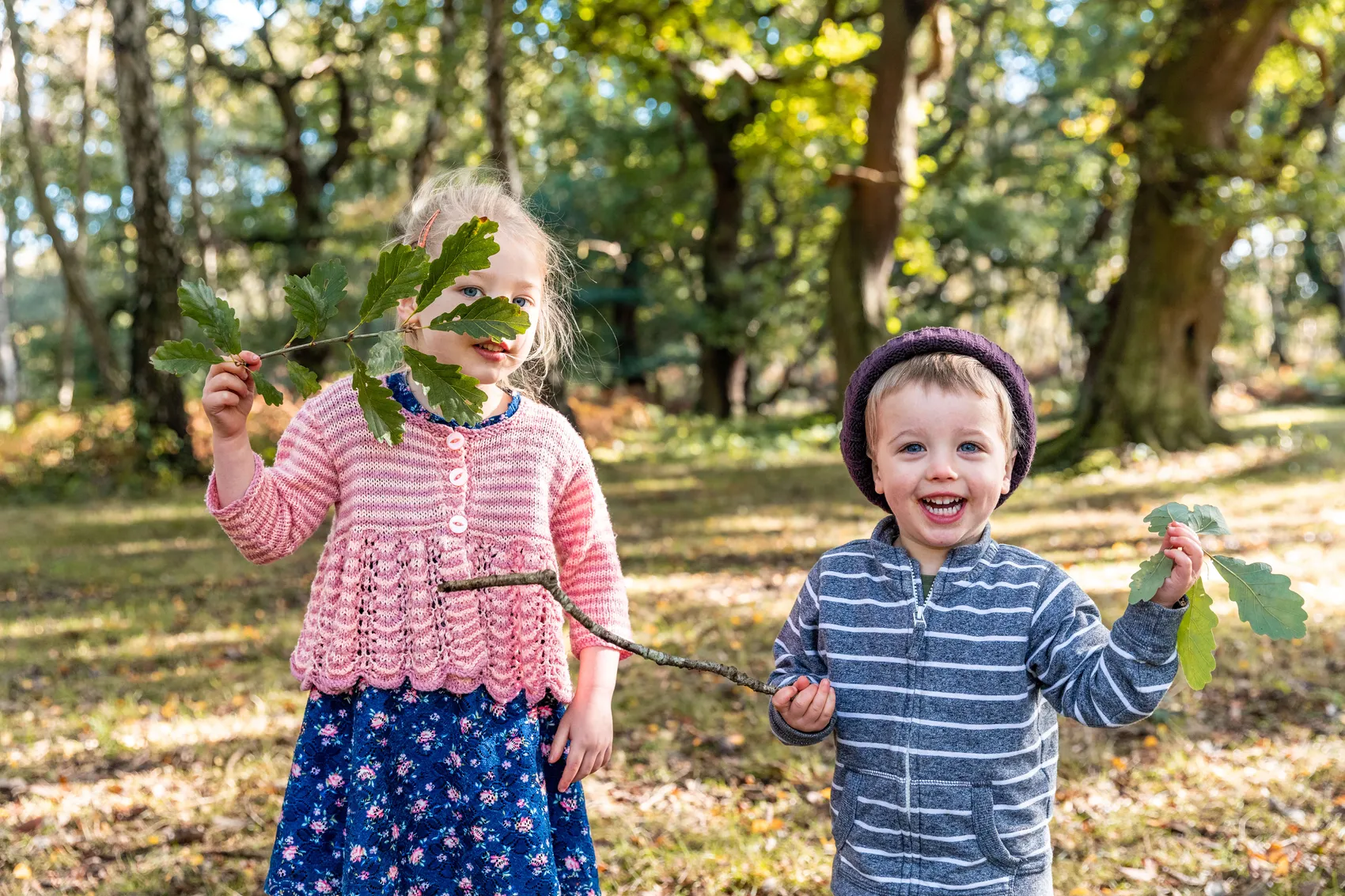 Zwei Kinder spielen im Wald: Ein Mädchen hält ein Eichenblatt vor sein Gesicht, ein Junge mit lila Mütze lächelt.