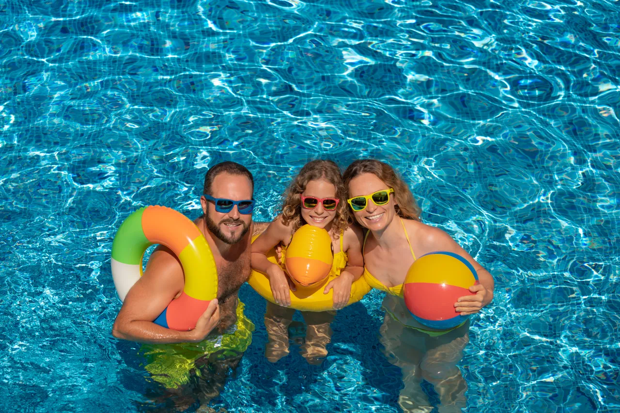 Eine Familie mit Sonnenbrillen steht in einem blauen Swimmingpool und hält bunte Schwimmreifen sowie einen Ball.