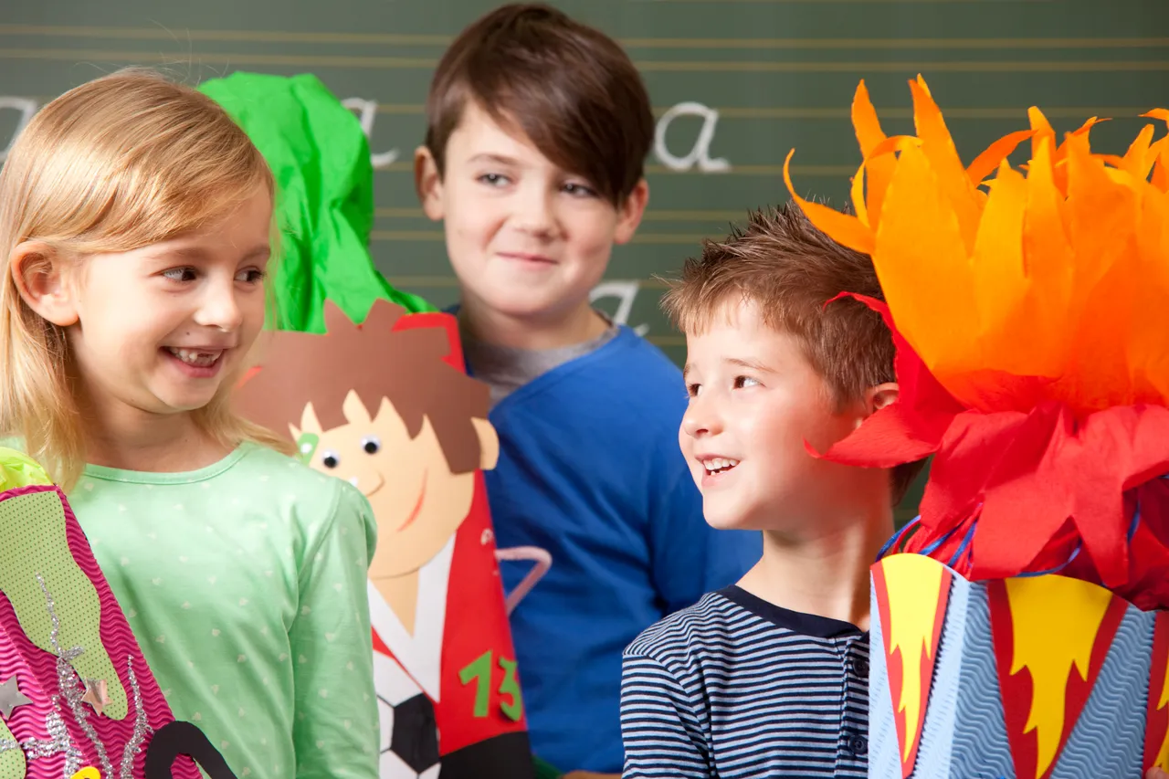 Drei lächelnde Kinder stehen in einem Klassenzimmer vor einer Tafel neben einer bunten Schultüte und einer Papierfigur.