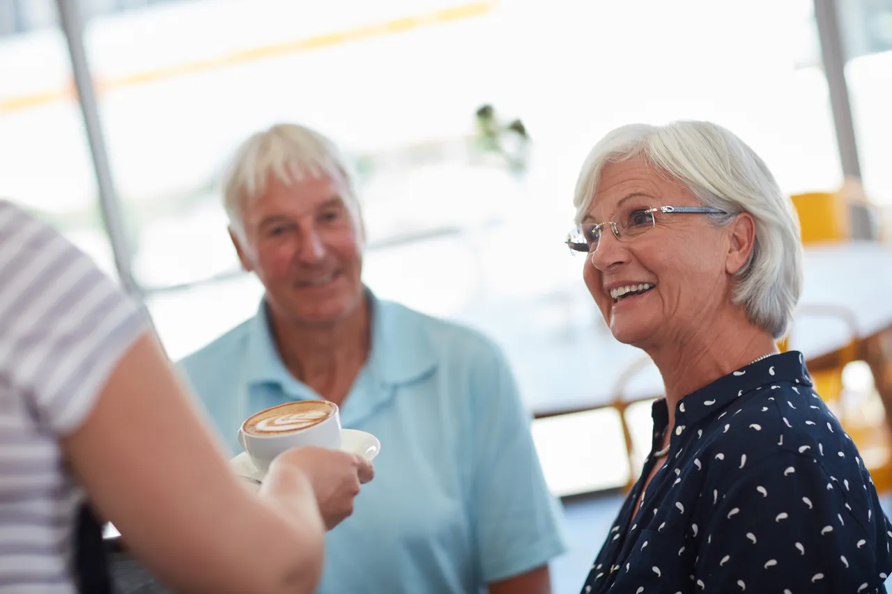 Eine lächelnde ältere Frau in einem Café, während ihr eine Tasse Kaffee serviert wird. Ein älterer Mann sitzt im Hintergrund.
