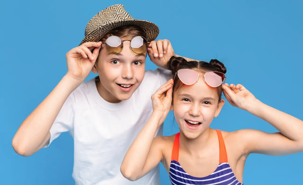 Zwei lächelnde Kinder setzen sich Sonnenbrillen auf. Der Junge trägt einen Strohhut, das Mädchen einen gestreiften Badeanzug.