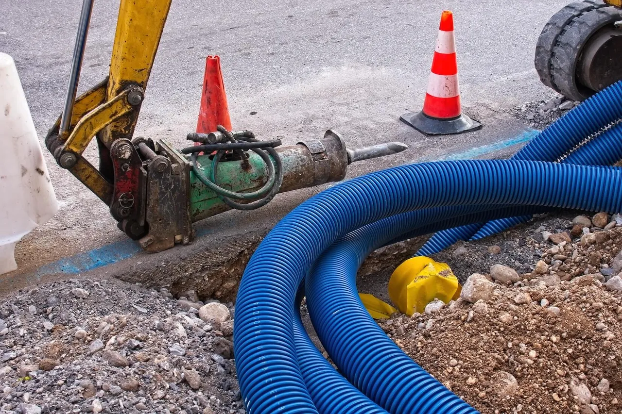 Ein gelber Bagger mit Abbruchhammer arbeitet neben hellblauen Wellrohren in einem Graben auf einer Baustelle.
