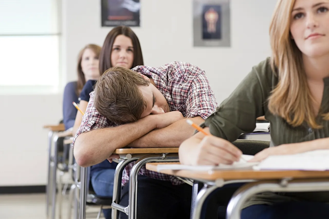 Ein Schüler schläft während des Unterrichts am Tisch, während eine Mitschülerin neben ihm schreibt und eine Lehrerin im Hintergrund aufmerksam beobachtet.