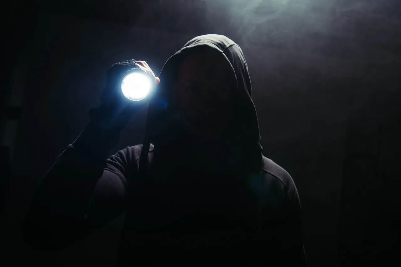 Eine Person mit Kapuze hält eine Taschenlampe in einem dunklen, verrauchten Raum.