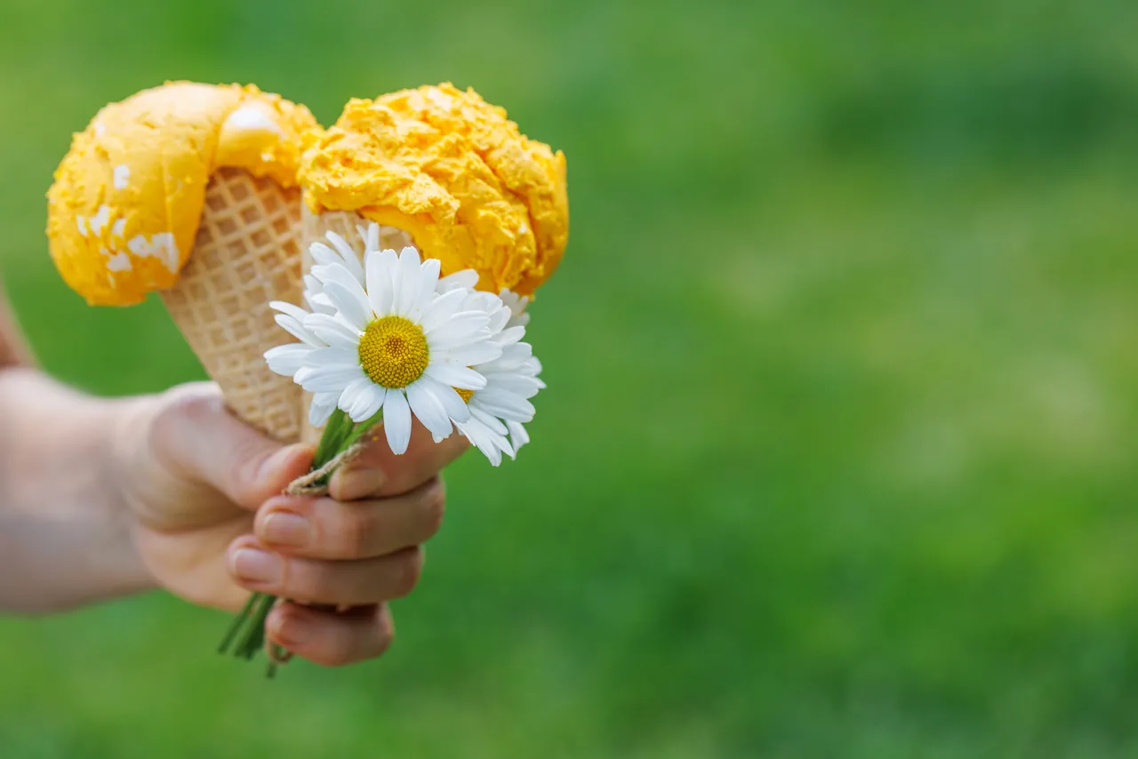 Zwei Waffeln mit gelbem Eis und eine weiße Gänseblümchen werden in einer Kinderhand gehalten. Der Hintergrund ist unscharf grün.