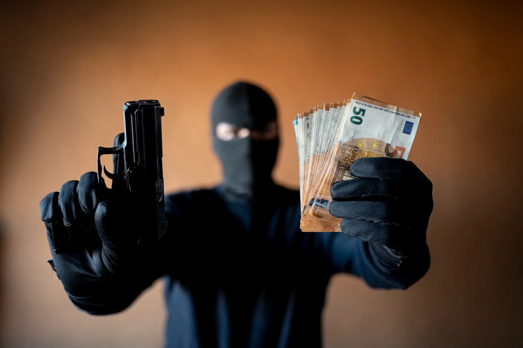 Person mit Sturmhaube hält eine Pistole und einen Fächer mit Geldscheinen in der Hand.
