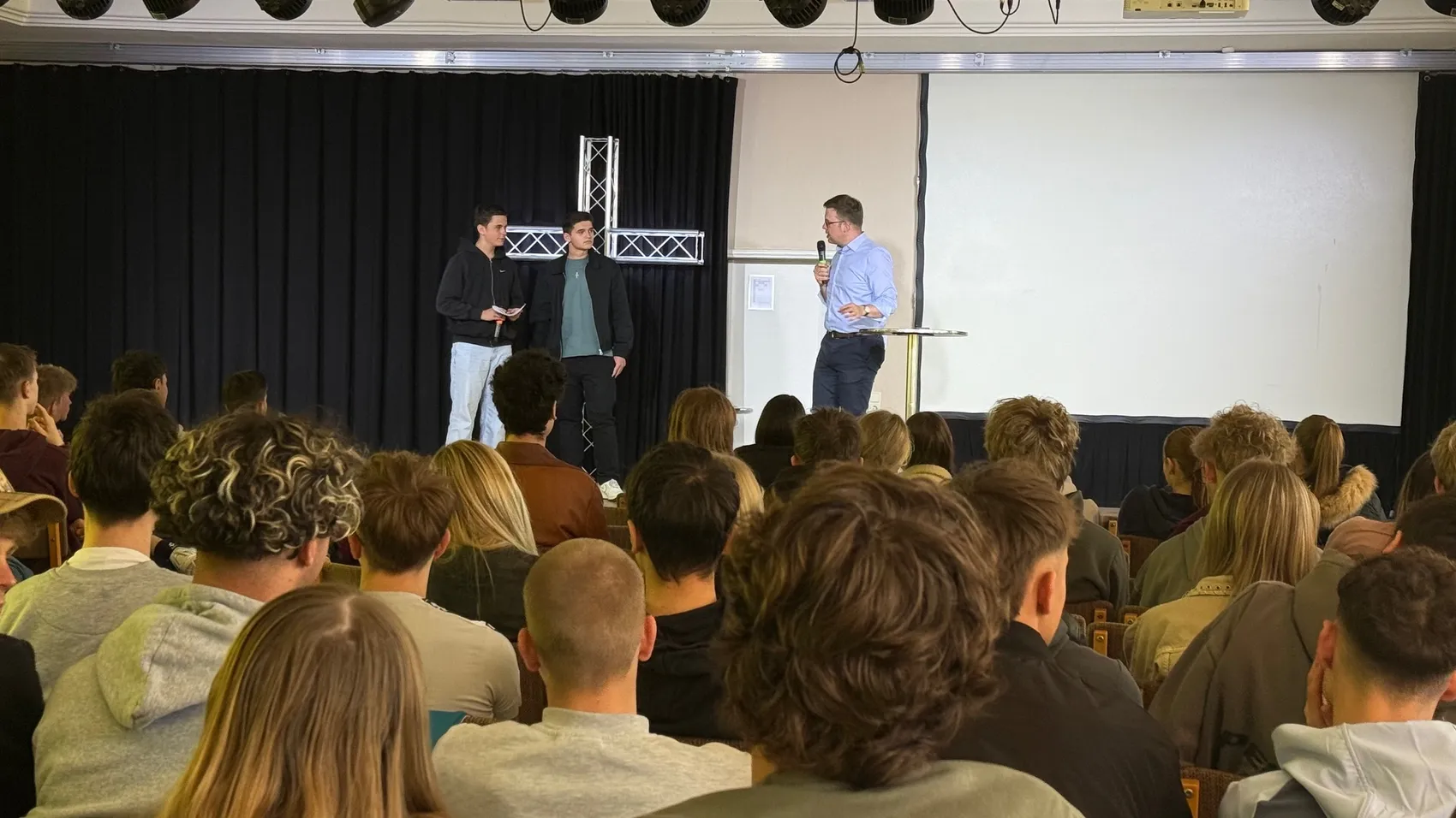 Florian Müller spricht vor einer Gruppe Schüler in einem Auditorium. Zwei weitere Personen stehen neben ihm auf der Bühne.