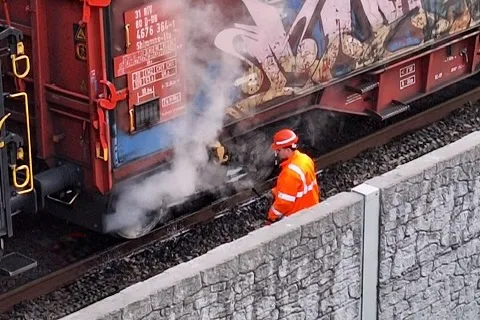 Ein Feuerwehrmann in orangefarbener Schutzkleidung und rotem Helm steht neben einem Güterwaggon, aus dem Rauch aufsteigt. Im Hintergrund ist eine Steinmauer zu sehen.