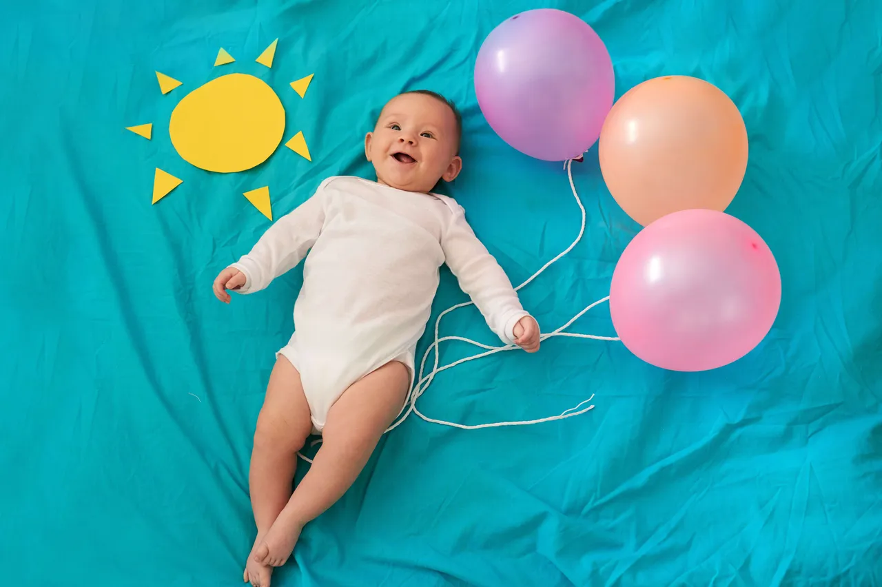Ein lächelndes Baby in einem weißen Body liegt auf einem blauen Tuch neben einer gelben Papier-Sonne und drei bunten Luftballons.