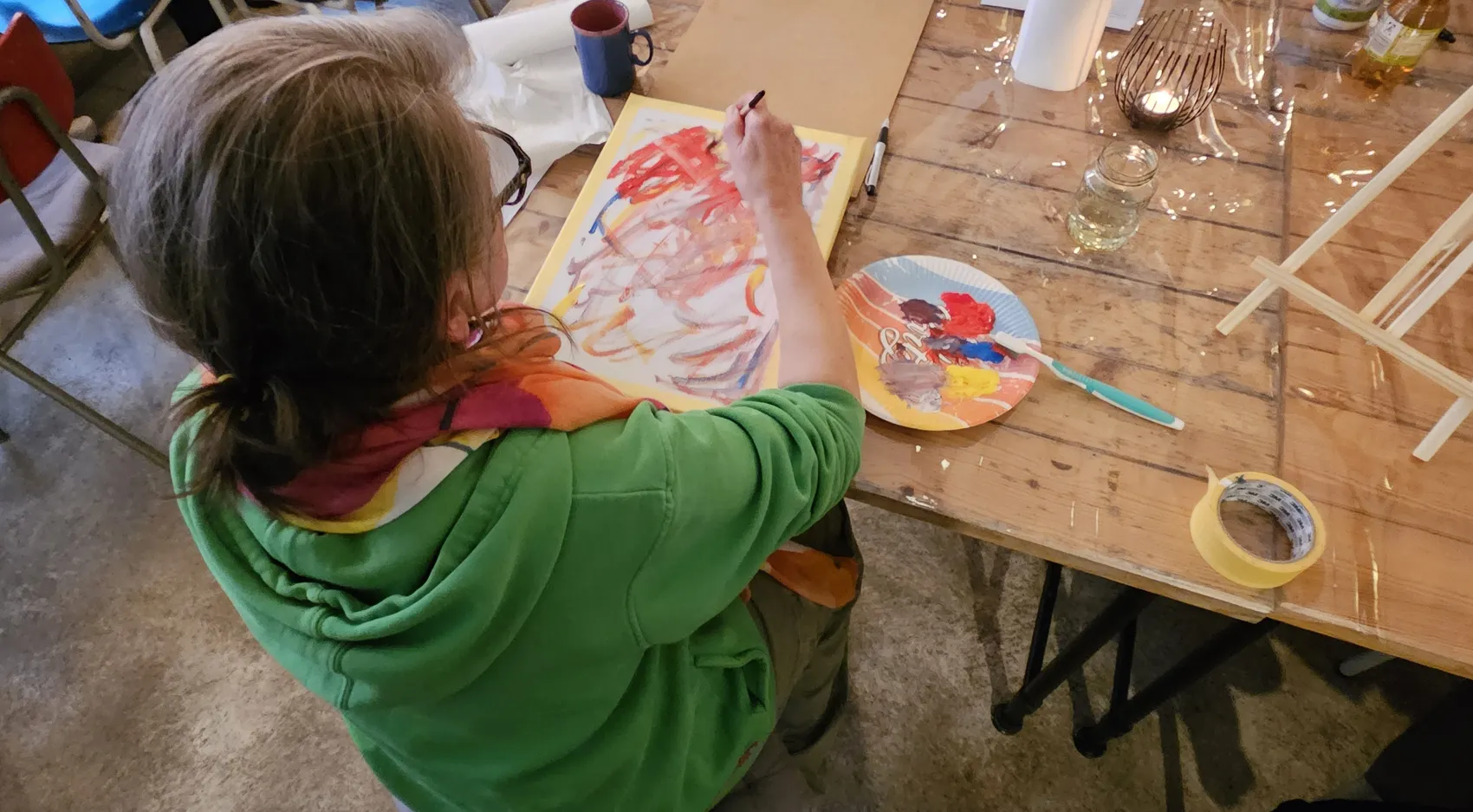 Eine Person malt mit Pinsel und Farben auf einem großen Papier, während auf einem Teller daneben weitere Farben bereitstehen. Der Fokus liegt auf dem kreativen Prozess des intuitiven Malens.