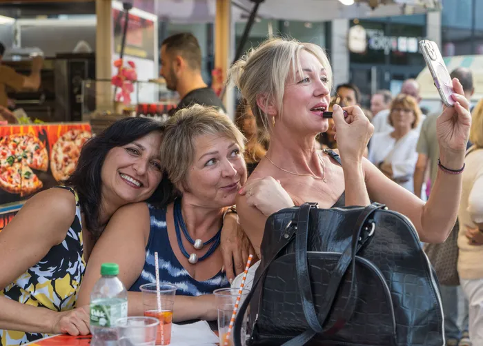 Drei Frauen lächeln gemeinsam, während eine von ihnen Lippenstift aufträgt und dabei in ihr Smartphone blickt.