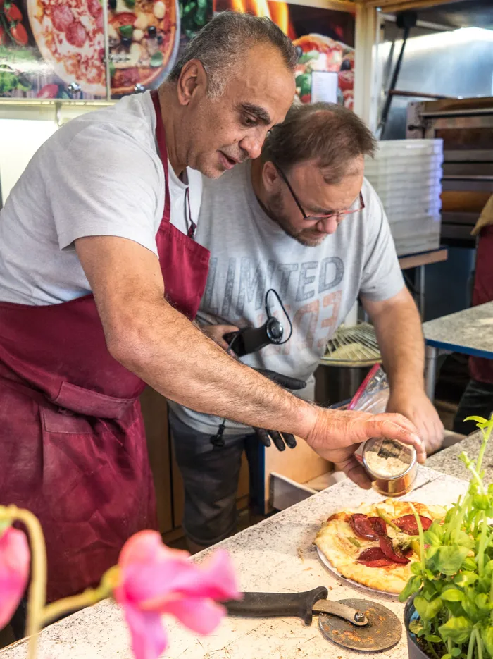 Ein Mann in weinroter Schürze bereitet an einem Verkaufsstand eine Pizza vor, während ein weiterer Mann daneben steht.