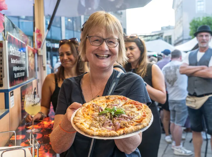 Eine lächelnde Frau mit Brille hält eine belegte Pizza auf einem weißen Teller in die Kamera.