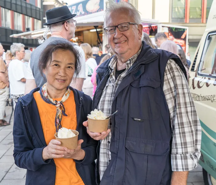 Ein Mann und eine Frau lächeln und halten kleine Becher mit weißem Dessert vor Menschen und Verkaufsständen.