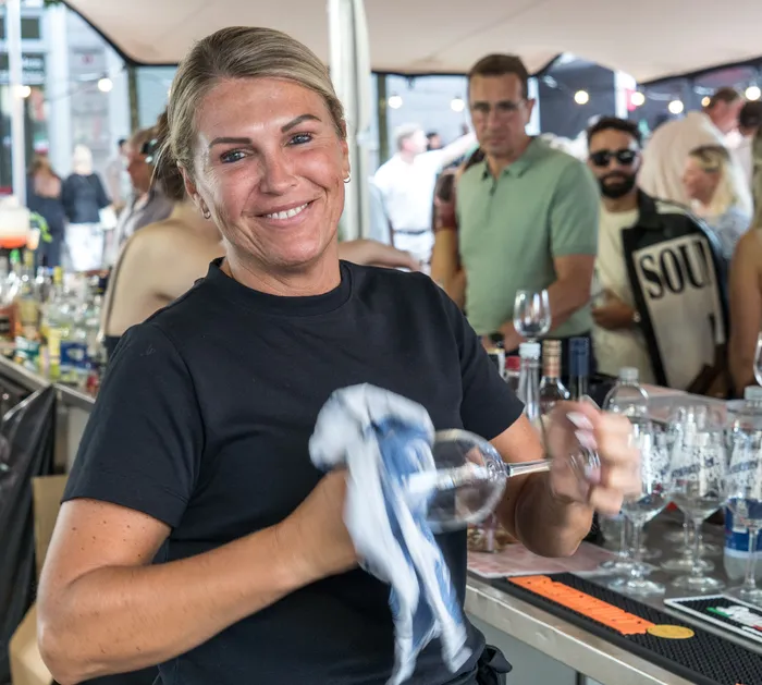 Eine lächelnde Frau in einem schwarzen T-Shirt poliert ein Weinglas an einer Bar.