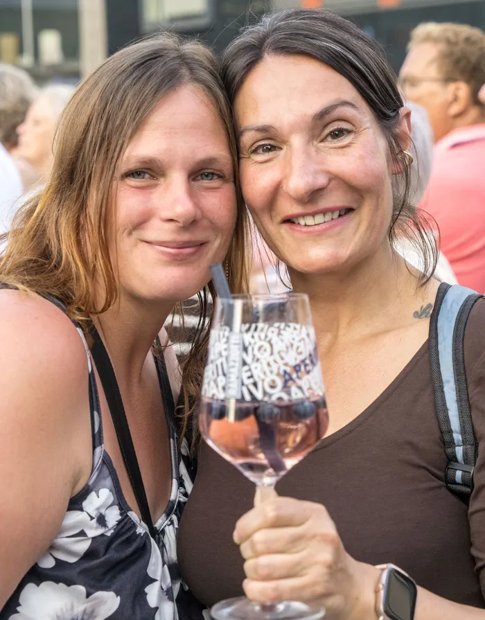 Zwei lächelnde Frauen posieren eng beieinander, eine hält ein Glas Roséwein mit einem Strohhalm.