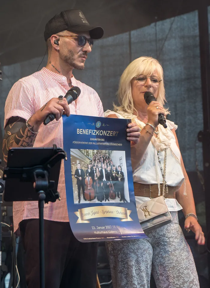 Ein Mann und eine Frau präsentieren ein Plakat für ein Benefizkonzert zugunsten des Fördervereins der Palliativstation Lüdenscheid.