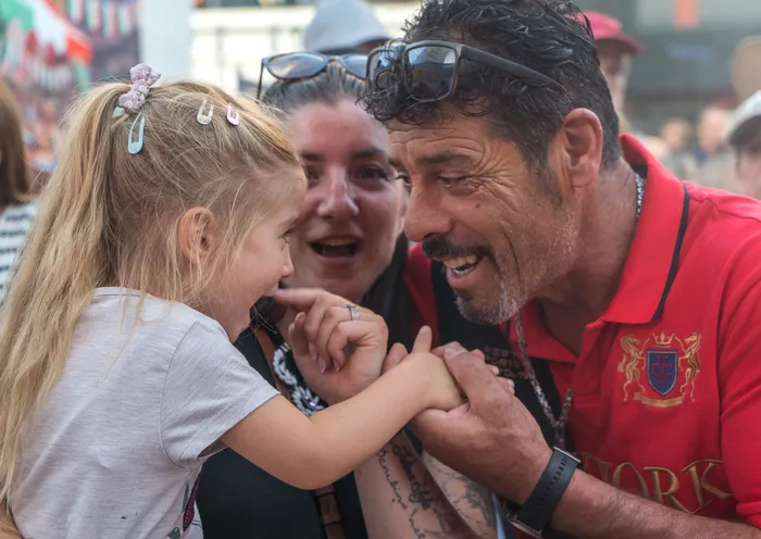 Ein lächelnder Mann in einem roten Poloshirt hält die Hand eines kleinen blonden Mädchens, während im Hintergrund eine Frau mitlächelt.