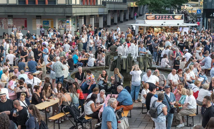 Eine große Menschenmenge sitzt an Tischen auf einem belebten Platz um einen Brunnen herum, im Hintergrund steht ein Pizza-Imbisswagen.