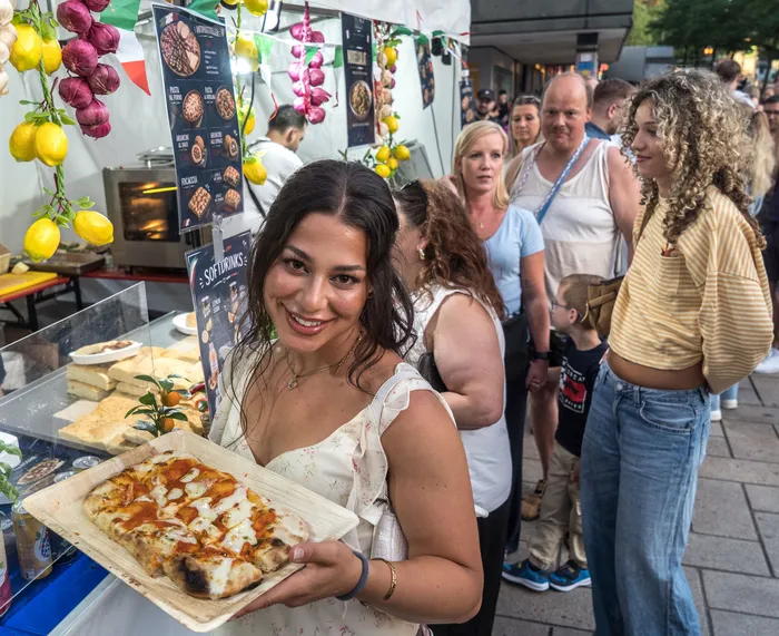 Eine lächelnde Frau hält ein Holztablett mit einer Pizza vor einem italienischen Essensstand bei der italienischen Nacht in Lüdenscheid.