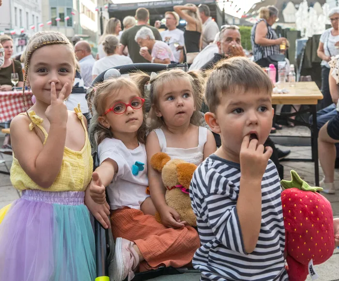 Vier Kinder posieren im Vordergrund bei einem belebten Open-Air-Event, darunter ein Mädchen in einem bunten Tutu und ein Junge im gestreiften Shirt.