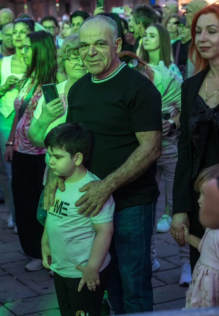 Ein Mann in einem schwarzen T-Shirt legt einem kleinen Jungen in einem weißen T-Shirt die Hände auf die Schultern inmitten einer Menschenmenge.