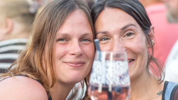 Zwei lächelnde Frauen halten ihre Köpfe eng aneinander, im Vordergrund ist ein Glas mit einem Getränk unscharf zu sehen.