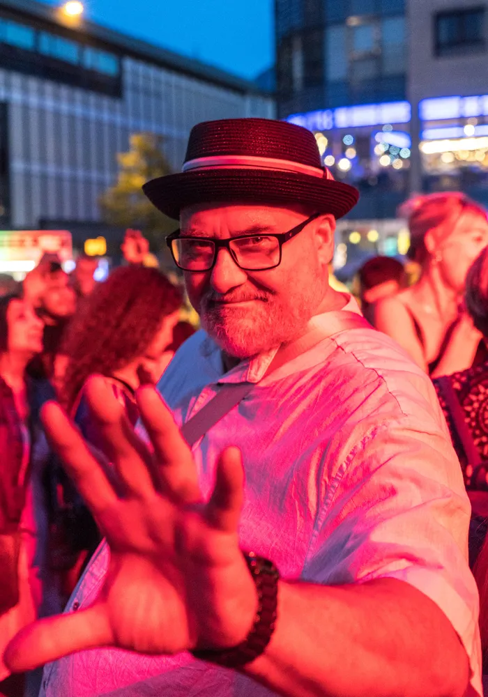 Ein bärtiger Mann mit Brille und schwarzem Hut streckt seine Hand in die Kamera vor einer Menschenmenge bei der italienischen Nacht in Lüdenscheid.